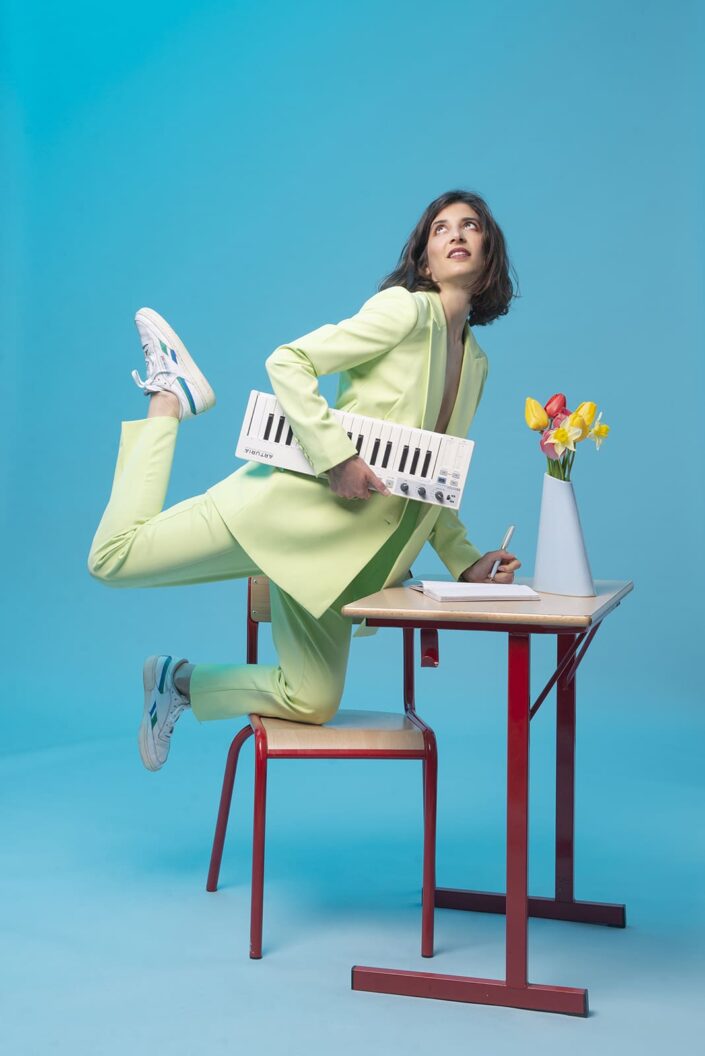 Photo portrait d'une femme avec une veste verte, sur une chaise avec un piano dans une main et de l'autre elle écrit dans un cahier sur un table d'école. Un vase de fleurs est posé sur la table. Elle regarde vers le haut